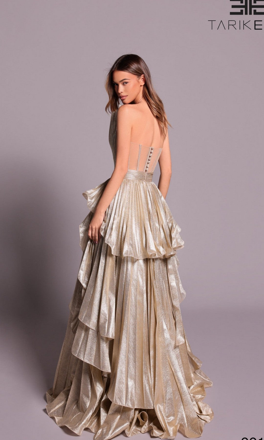 A woman models the Tarik Ediz 99148 long formal dress—a shimmery gold, strapless gown with layered ruffles and a semi-sheer back—posed against a plain light grey background.
